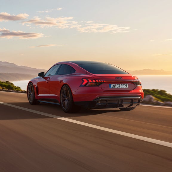 Der e-tron GT quattro auf einer kurvigen Küstenstraße bei Sonnenschein, zur Rechten das Meer, zur Linken felsiges Gebirge. 