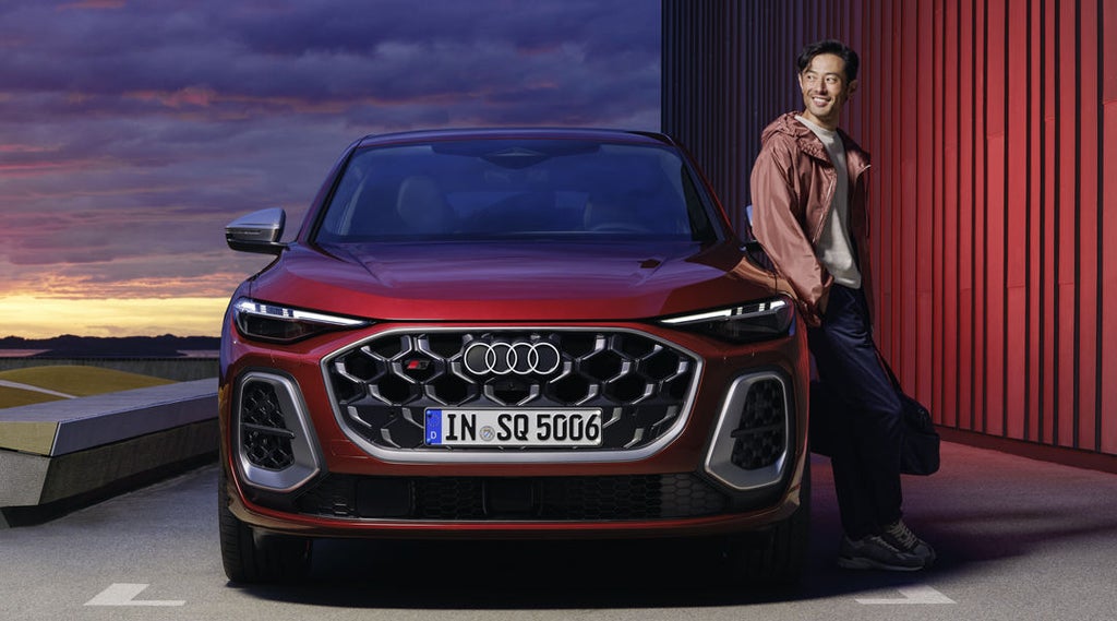 Der rote SQ5 Sportback steht auf einem Parkplatz parallel zu einem Hafen. Ein junger Mann lehnt sich an das Fahrzeug.