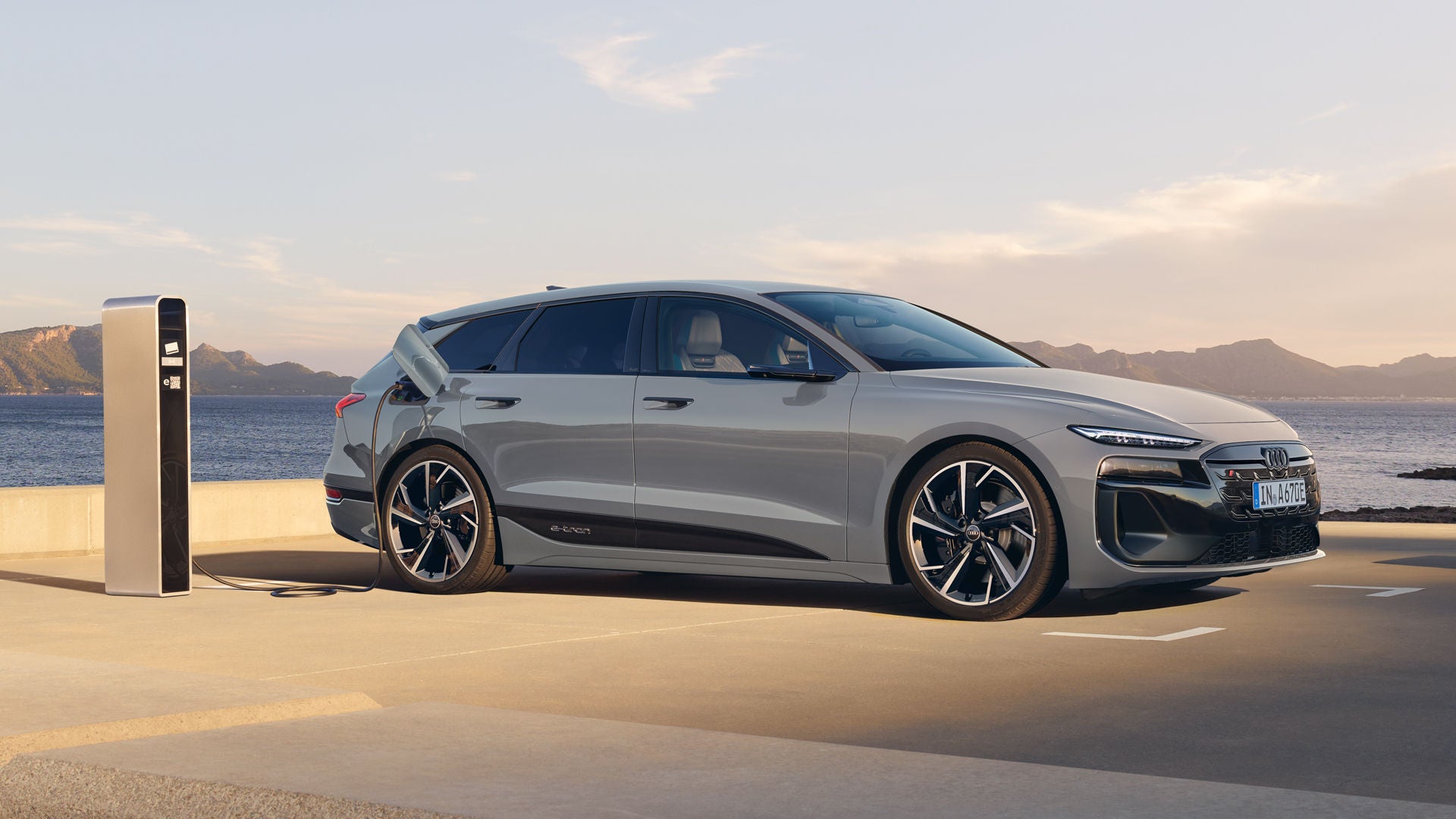 Der Audi S6 Avant e-tron mit mit Individuallackierung Audi exclusive Pfeilgrau Perleffekt, leicht schräg von der Seite fotografiert, auf einem Parkplatz direkt an der Küste. Am Horizont sind Gebirge zu erkennen.