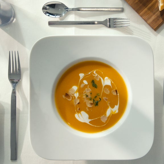 Obenansicht eines Tellers mit Kürbissuppe, garniert mit Sahne, Kräutern und Mandeln, umgeben von Besteck auf weißer Tischdecke.