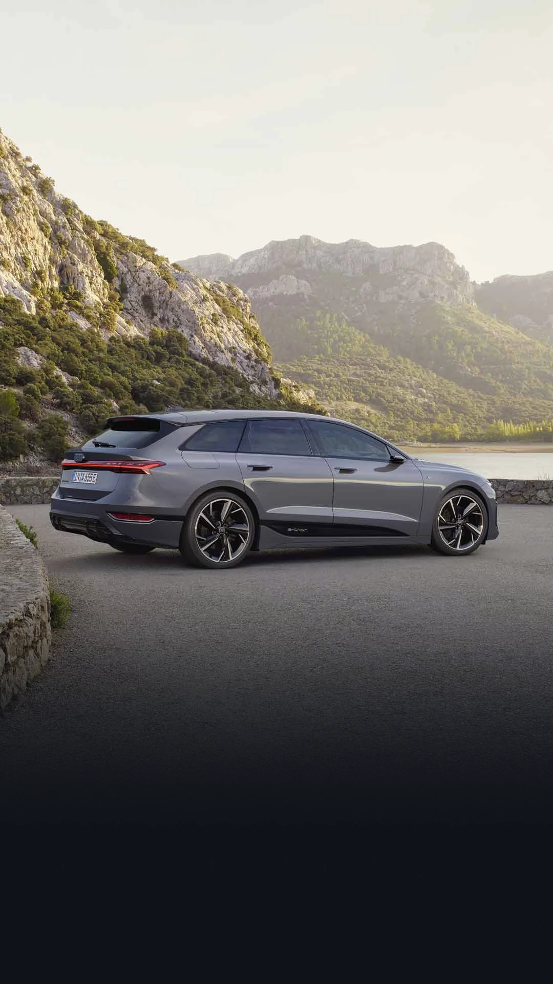 The A6 Avant e-tron is parked sideways on a gravel car park in front of a lake. Tree-covered mountains can be seen in the background.