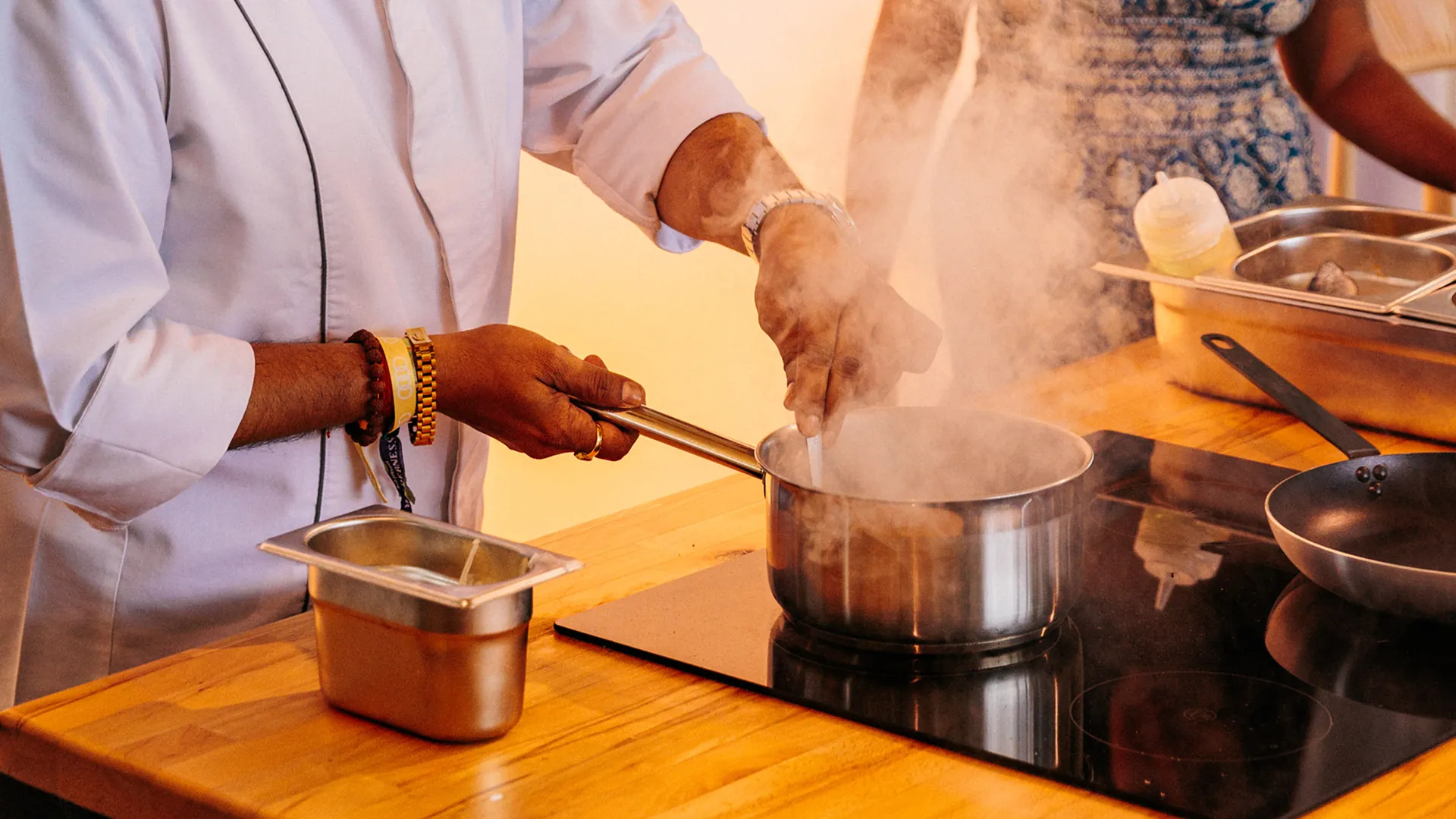 Close-up of a chef's hands stirring a steaming pot on an induction hob, with prep trays and a wok on the wooden counter beside it. Close-up of a chef's hands stirring a steaming pot on an induction hob, with prep trays and a wok on the wooden counter beside it.