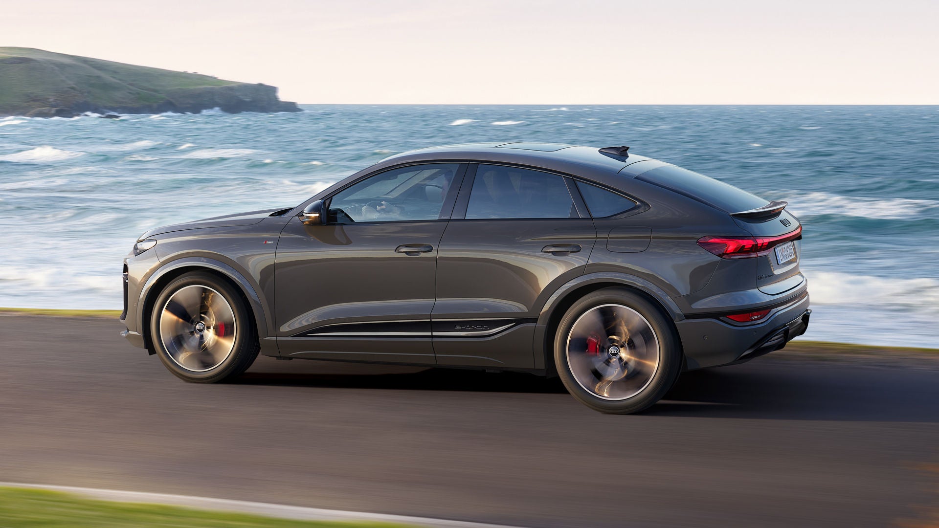 Seitliche Aufnahme des Q6 Sportback e-tron, welcher auf einer Straße entlang des Meeres fährt.