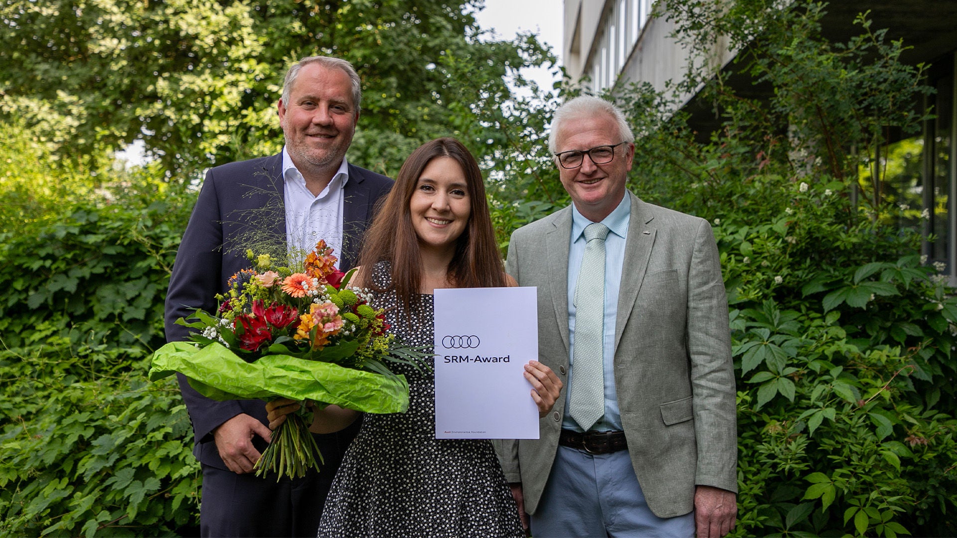 Drei Personen stehen lächelnd vor grüner Kulisse; eine Frau in der Mitte hält Blumen und ein Dokument mit der Aufschrift „Audi SRM-Award“.