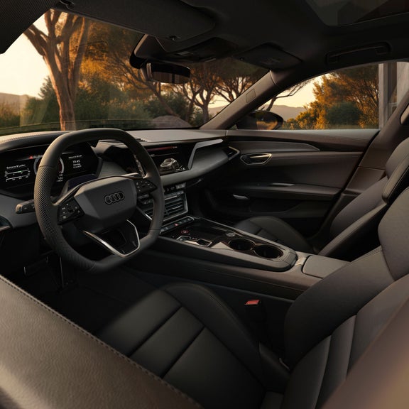 Der Innenraum eines Audi e-tron GT quattro mit Blick in einen Garten. Zu erkennen sind Sitze, das Lenkrad und die Amaturen. Die Szenerie ist in das Licht eines Sonnenuntergangs gehüllt.