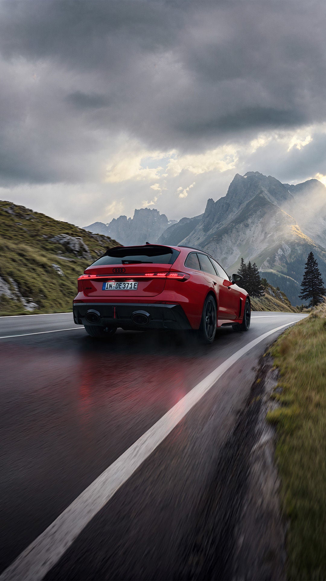 Red Audi RS 5 Avant wagon driving on a winding mountain road, dramatic Alpine backdrop with clouds and sunbeams in the background.