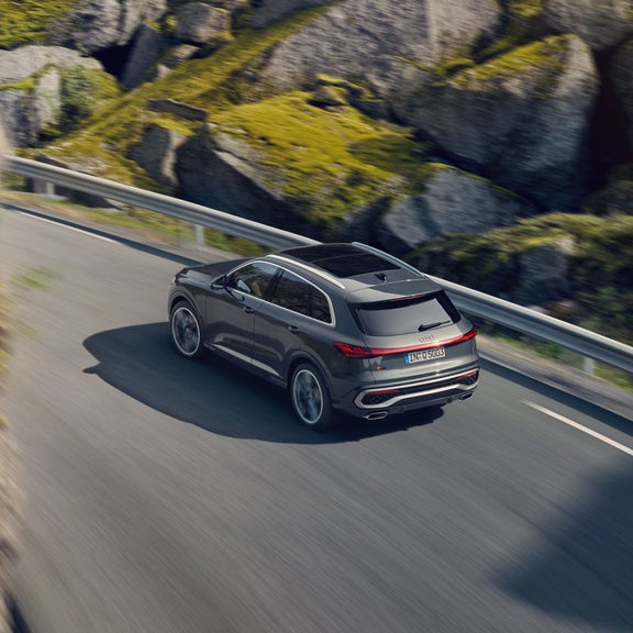 A grey SUV driving along a curved mountain road with rocky scenery.