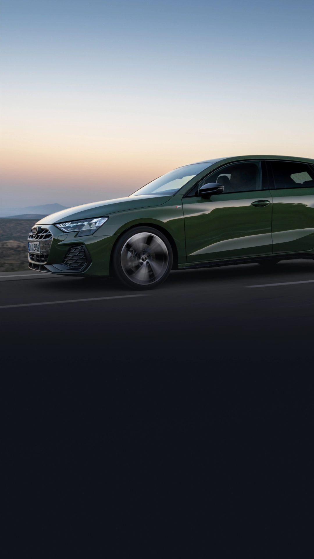 Green Audi A3 Sportback driving on road, front left side visible, against sunset backdrop.