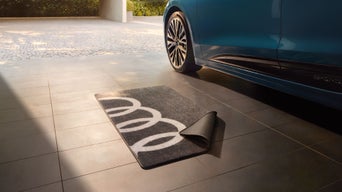 Teppich mit Audi-Ringen auf Fliesenboden neben Fahrzeugreifen.