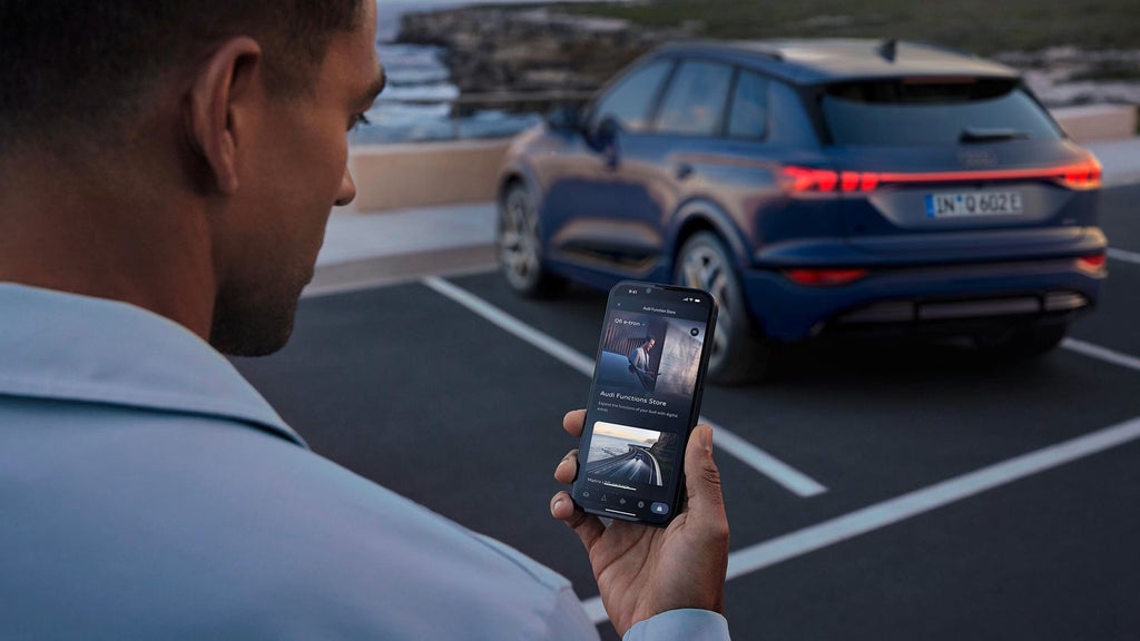 A person holds a smartphone displaying the Audi Functions Store, featuring parking assist options for an Audi vehicle.