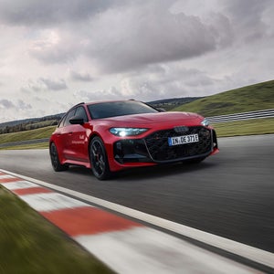 Ein roter Audi RS 5 Avant fährt auf einer Rennstrecke. Im Vordergrund sind rot-weiße Curbs zu erkennen, im Hintergrund eine grüne Hügellandschaft.