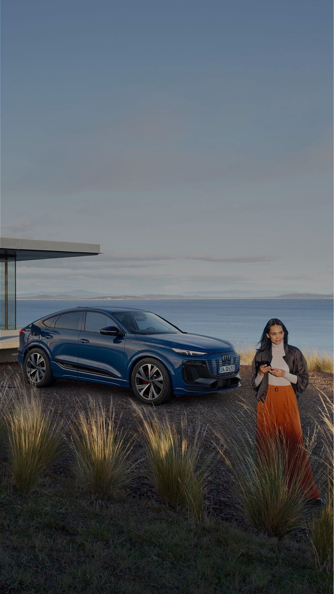A woman stands using her phone near a blue electric Audi SUV parked by a modern house and charging station, with a scenic coastal view in the background.