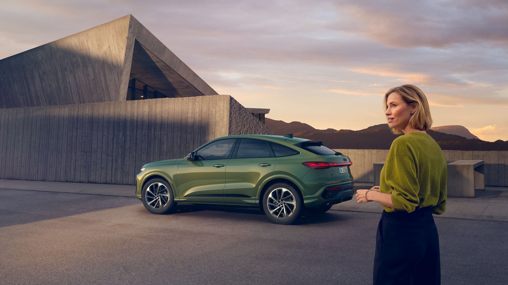 Eine Frau steht vor einem modernen Gebäude, im Hintergrund ein grüner Audi SUV und eine Berglandschaft bei Sonnenuntergang.