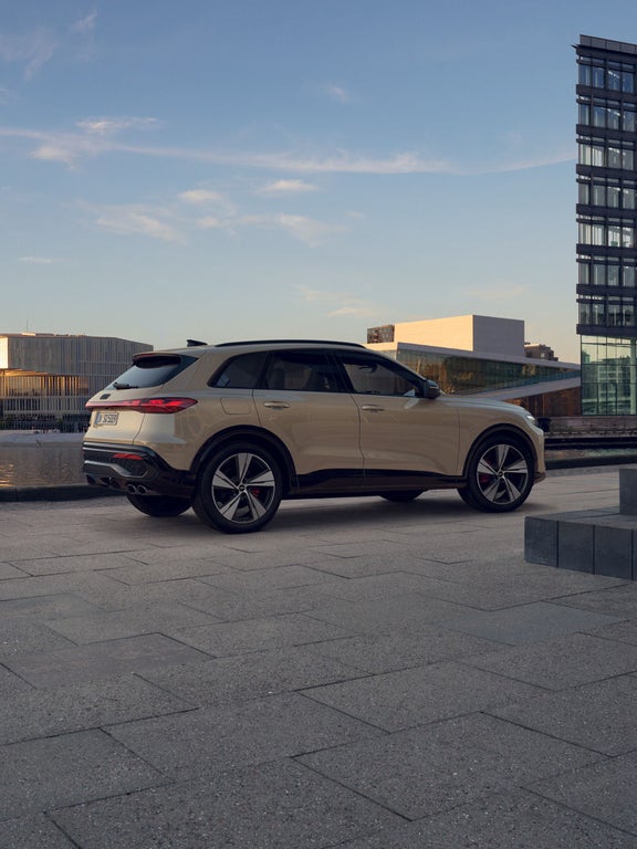 Ein beiger Audi SQ5 SUV von hinten seitlich, geparkt vor moderner Architektur bei Sonnenuntergang.
