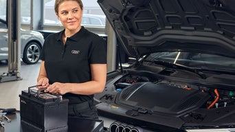 Frau hält eine Auto-Batterie und steht vor einem Audi mit offener Motorhaube
