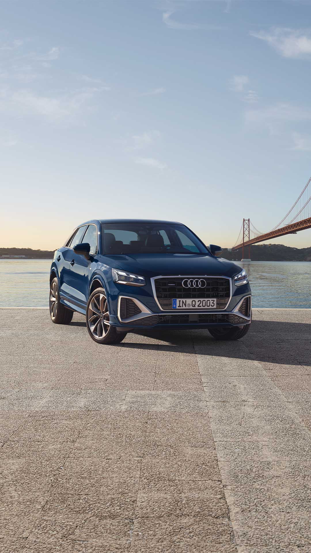 Ein blauer Audi Q2 von vorne vor Wasserlandschaft bei Sonnenaufgang, rechts im Bild eine rote Hängebrücke.