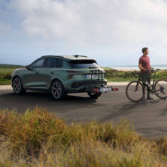 The Q3 with bike rack stands in front of a coastal landscape, next to it a young man with a gravel bike.