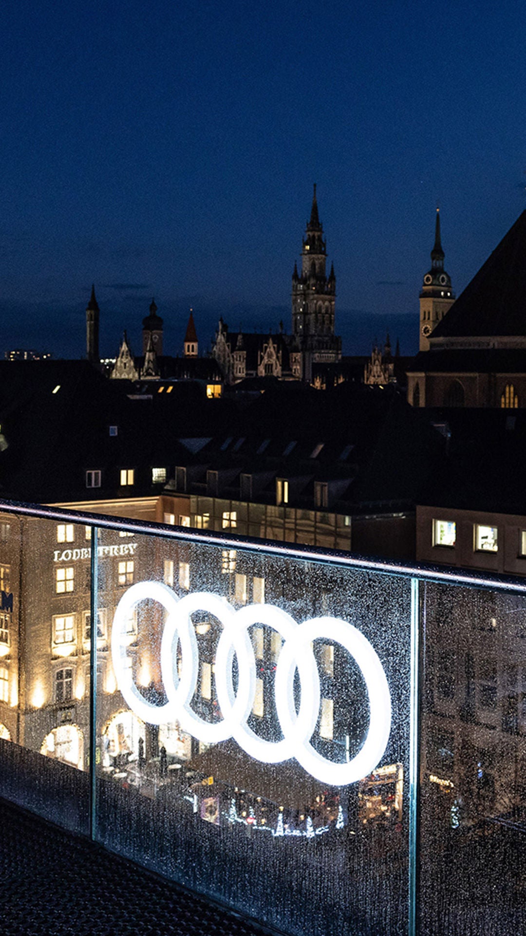 Blick über München bei Nacht mit beleuchtetem Audi-Logo im Vordergrund und Frauenkirche im Hintergrund.