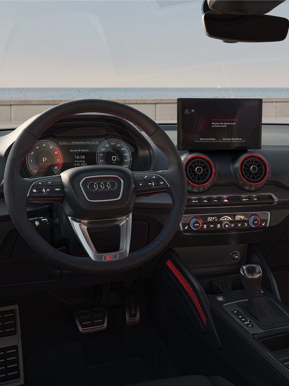 Innenraum eines Audi SQ2 mit Sportlenkrad und digitalen Anzeigen, Blick auf Bäume und Mauer durch die Frontscheibe.