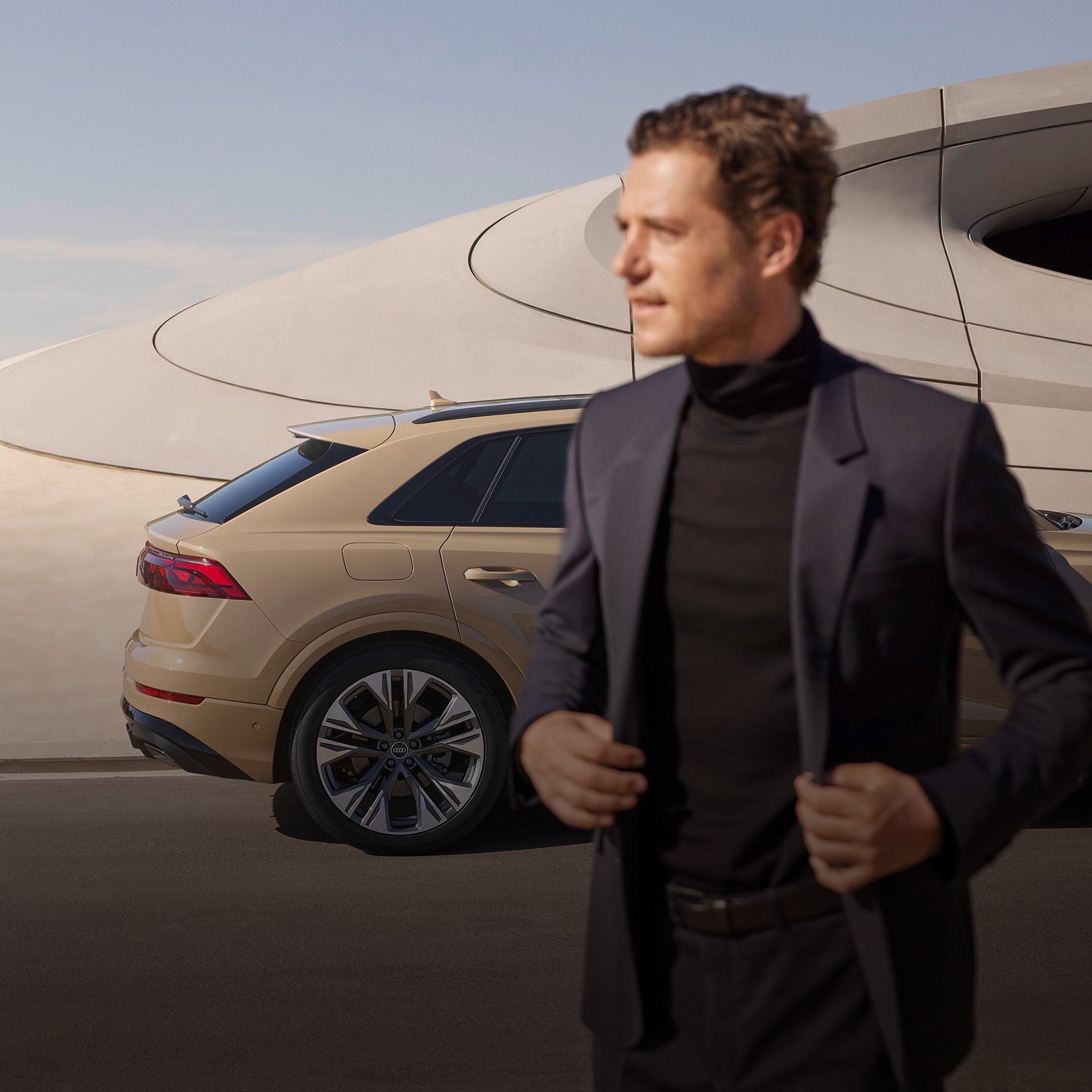 Ein stilvoll gekleideter Mann im dunklen Anzug steht unscharf im Vordergrund, während im Fokus ein goldener Audi Q8 SUV geparkt ist. Das Auto steht vor einem futuristischen, sandfarbenen Gebäude mit organischer Architektur. Der Himmel ist klar und blau – die Szene vermittelt Eleganz und urbanen Lifestyle.