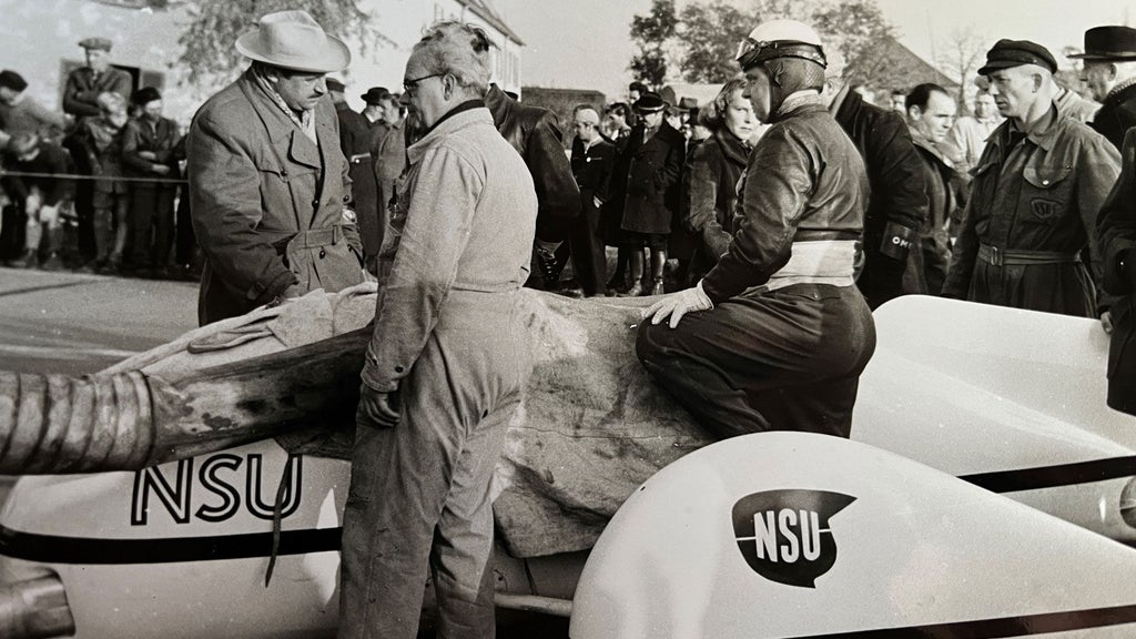 Black and white image of many people standing in front of a vehicle. One person is wearing a helmet