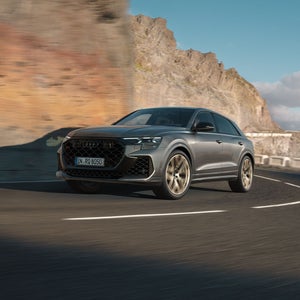Der RS Q8 SUV schräg von vorne in einer Kurve auf einer Bergstraße. Das Fahrzeug wirft Schatten an die rechts aufragende Bergwand.