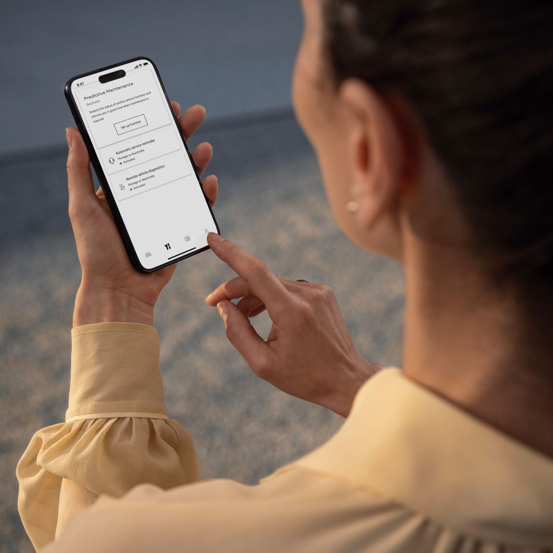 A woman in a yellow blouse holds a smartphone displaying a predictive maintenance app interface. She is tapping the screen, focused.