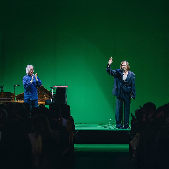 Zwei Männer stehen auf einer Bühne mit grünem Hintergrund; einer winkt, der andere applaudiert, Publikum im Vordergrund.