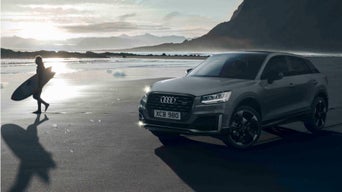 A beach in the late afternoon with a surfer walking on the left & a parked Audi vehicle on the right