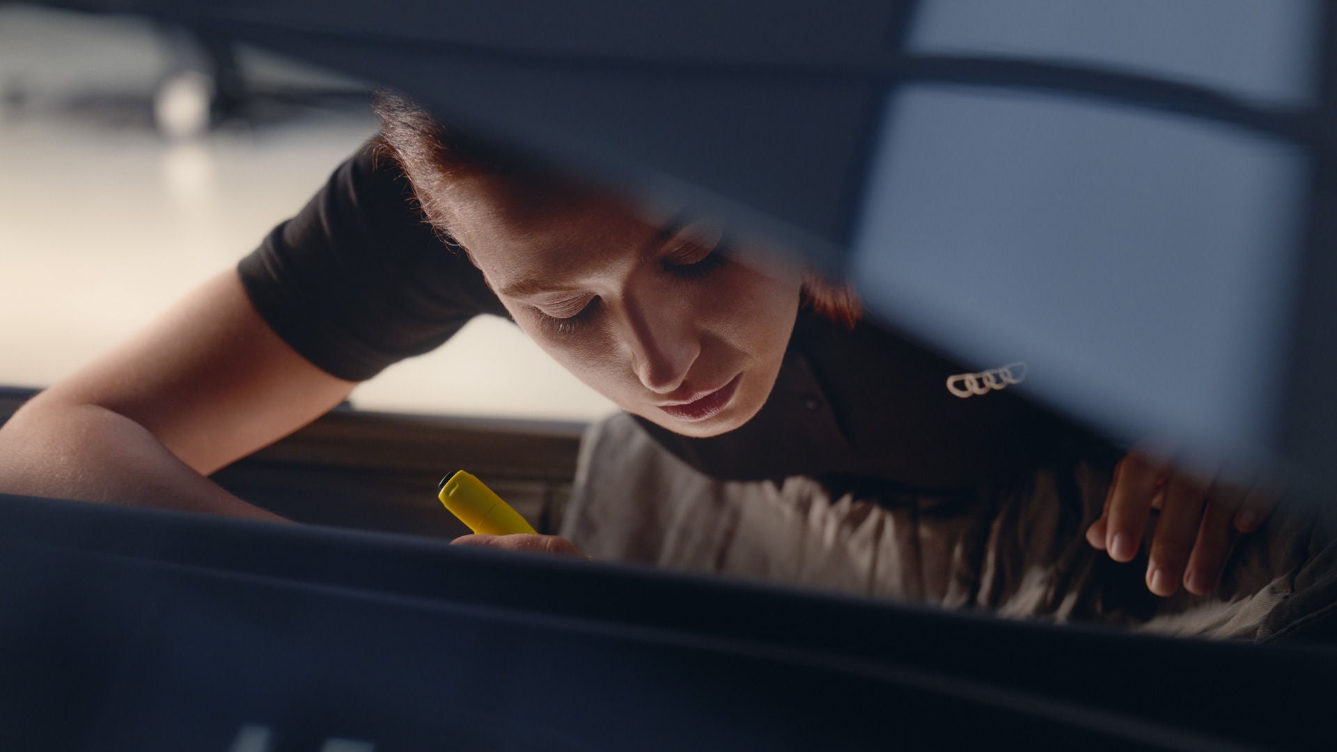 Eine Frau beugt sich über den Innenraum eines Fahrzeugs und hält einen gelben Gegenstand in der rechten Hand. Auf ihrer schwarzen Kleidung ist ein Audi-Logo zu sehen.   