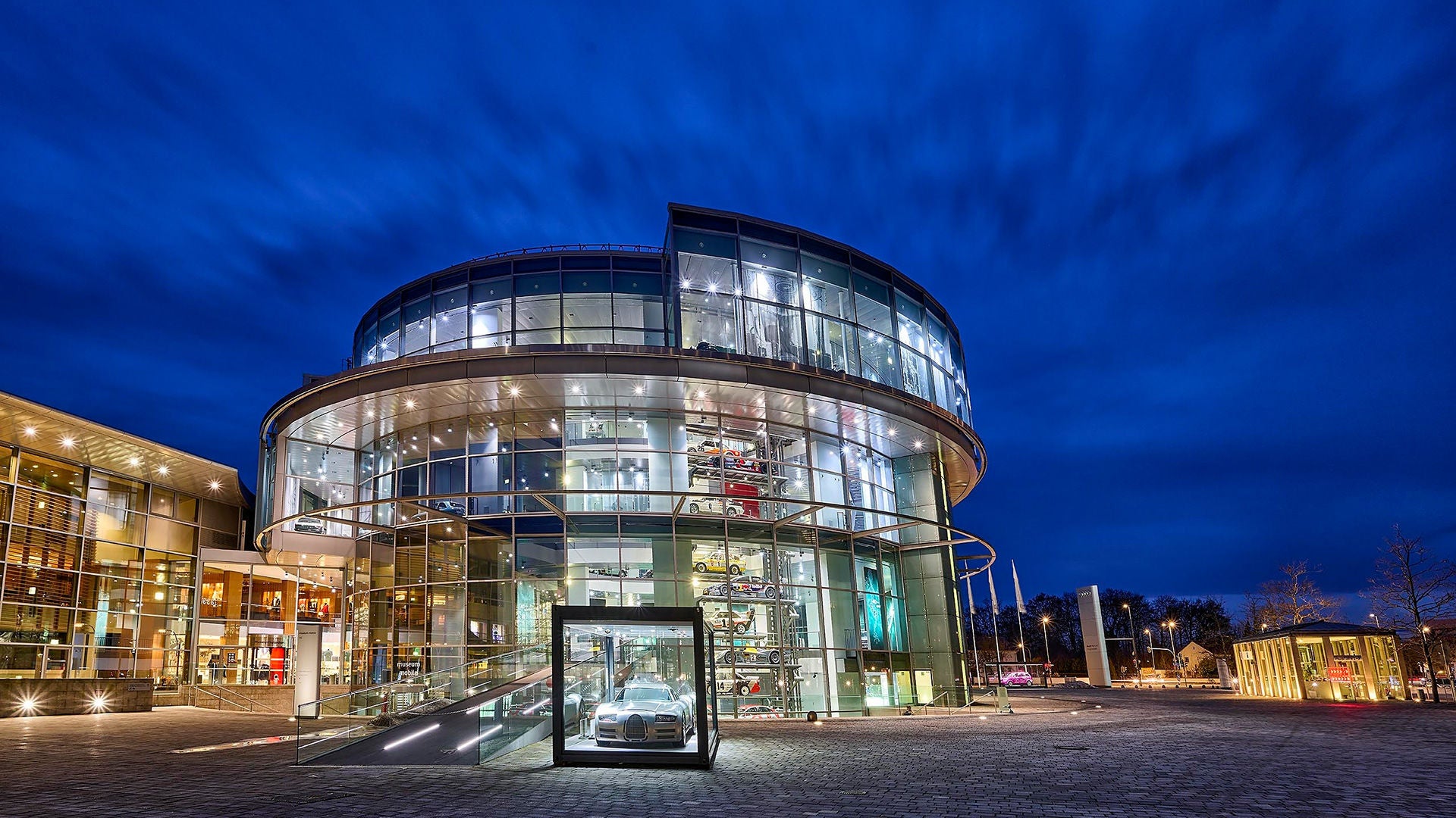 Modern, round glass building of the Audi Forum at night, blue illuminated sky in the background.