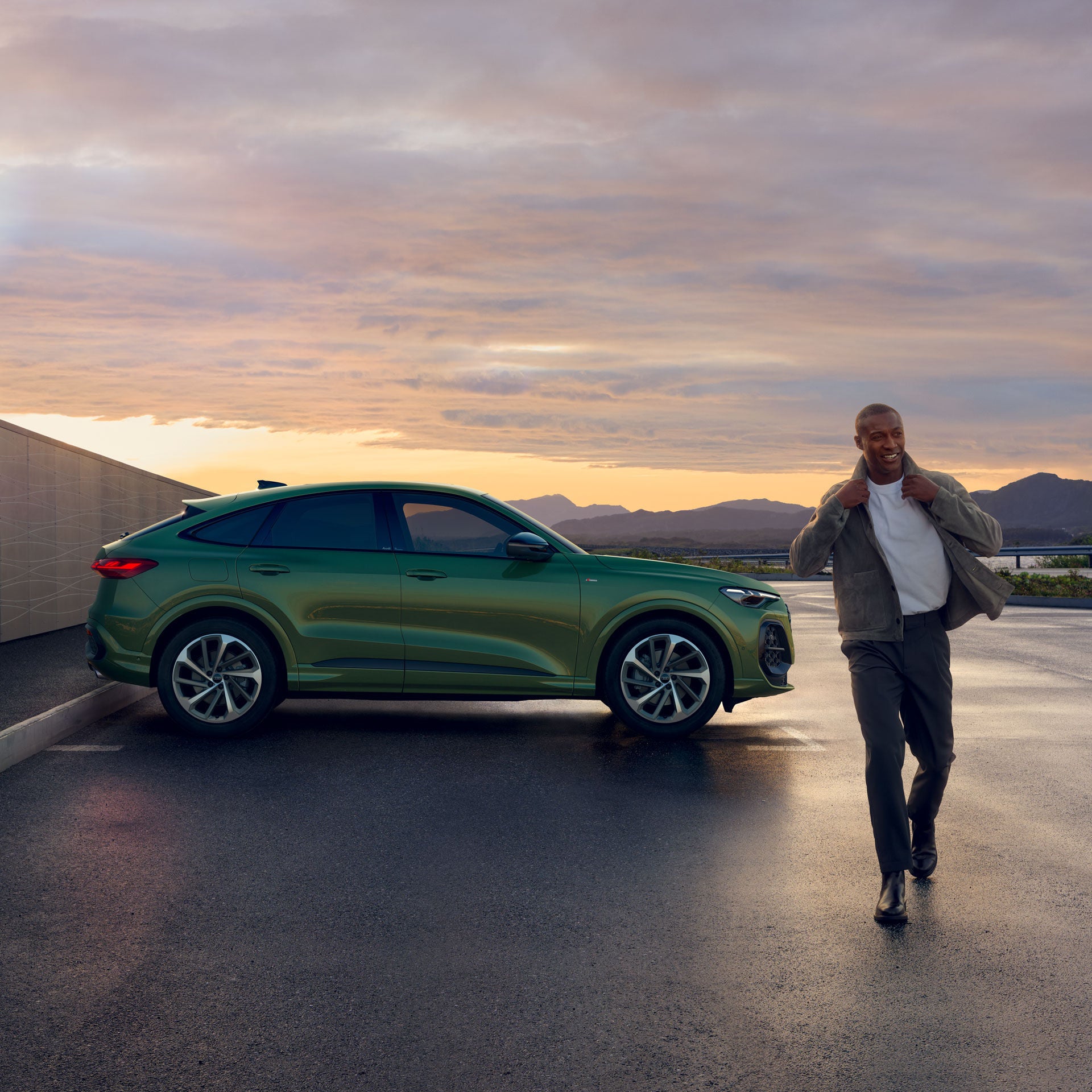 Mann geht lächelnd von einem grünen Audi Q5 bei Sonnenuntergang weg; im Hintergrund Berge und bewölkter Himmel.