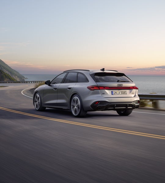 Der A5 Avant in einer schrägen Heckperspektive, während er bei Sonnenuntergang eine kurvige Küstenstraße entlangfährt.