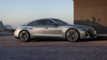 Ein silberner Audi S e-tron GT steht auf einer gepflasterten Auffahrt und ist an eine Ladestation angeschlossen. Im Hintergrund sind moderne Häuser und eine Dünenlandschaft zu sehen.
