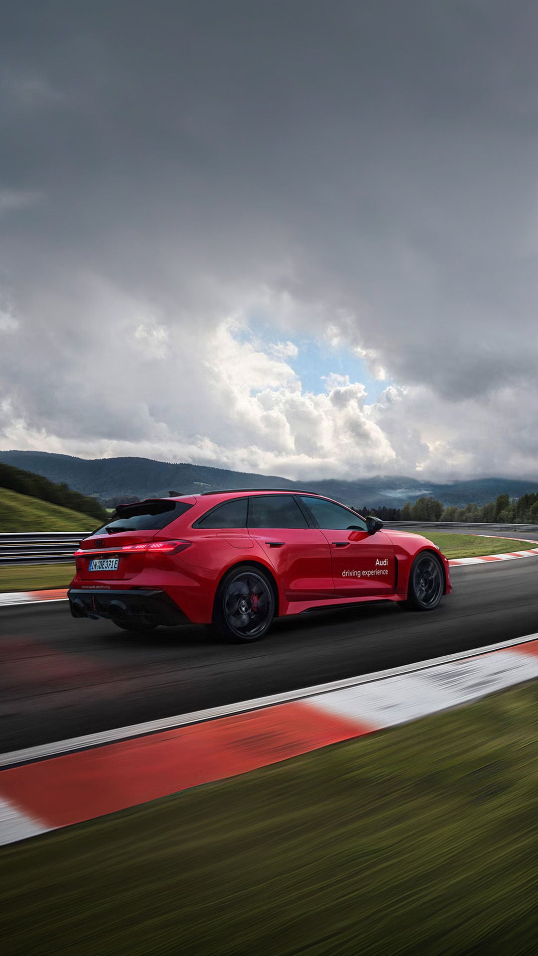 Roter Audi RS 5 Avant fährt durch eine Kurve auf der Rennstrecke; Leitplanken, grüne Hügel und wolkiger Himmel.
