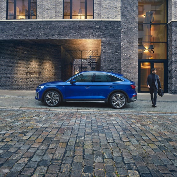 A blue SUV parked on a cobblestone street by a brick building with lit signage.