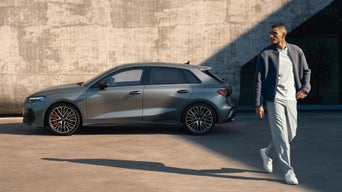 Mann geht vor einem grauen Audi S3 Sportback, der seitlich vor einer Betonwand im Sonnenlicht geparkt ist.