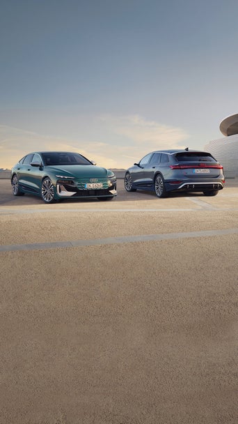 Two sleek Audi cars, a green sedan and a blue SUV, are parked on a concrete surface at dusk. The scene conveys modernity and sophistication.