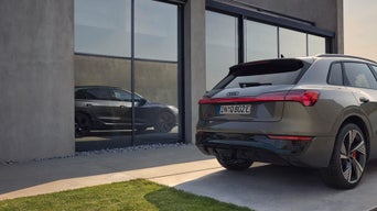 Grauer Audi-SUV vor modernem Gebäude; Rückansicht mit rotem Lichtband und Spiegelung in Glasfront.