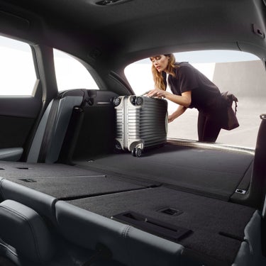 Frau lädt silbernen Koffer in den flachen, erweiterten Kofferraum eines Fahrzeugs mit umgeklappter Rücksitzbank.