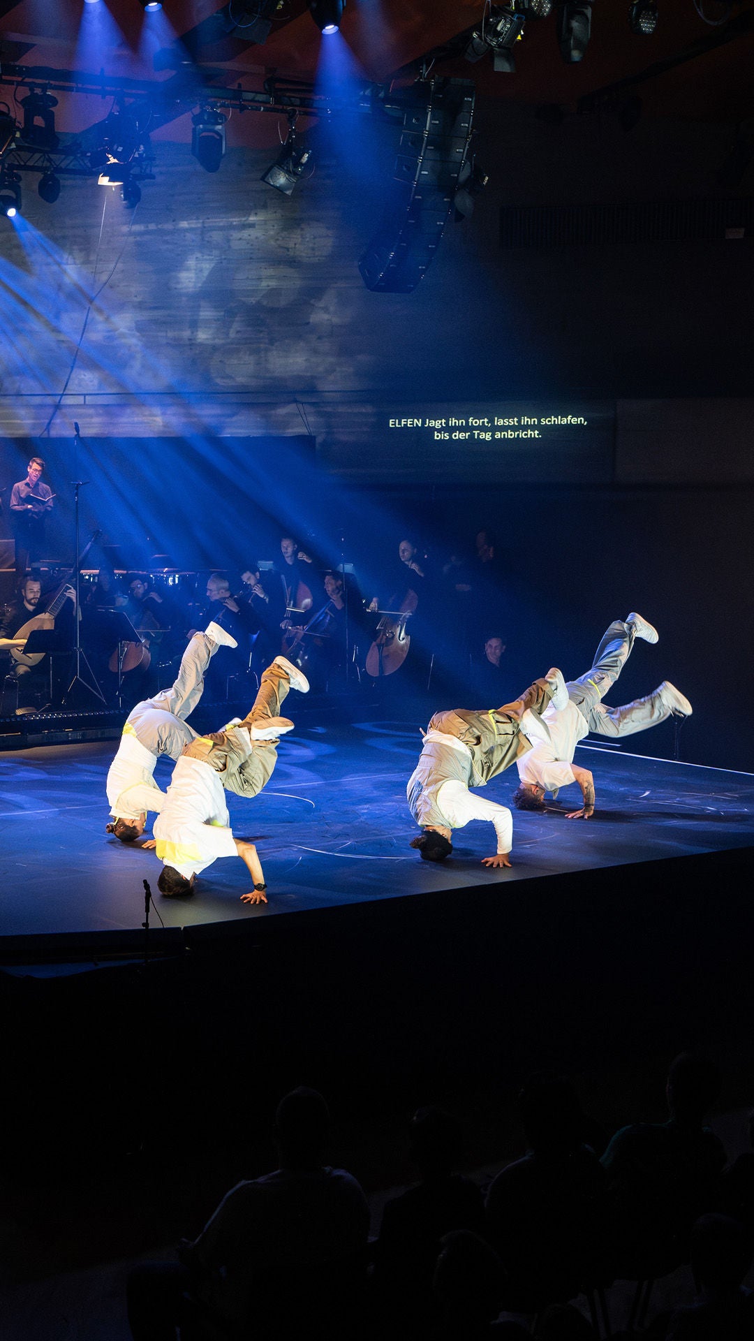 Vier Tänzer in weißer Kleidung bewegen sich auf der Bühne vor Orchester und Chor, blaues Bühnenlicht.