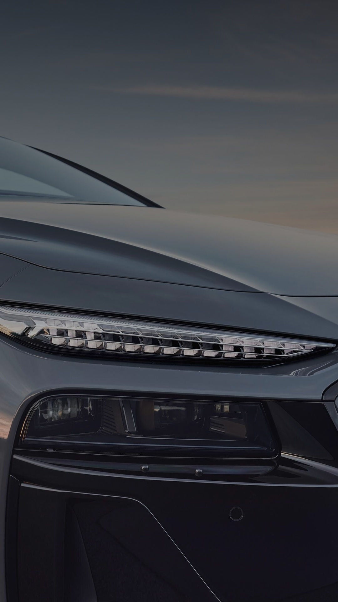 Close-up of an Audi car's front, highlighting a modern, angular headlight with LED elements. The grey surface reflects a subtle sunset.