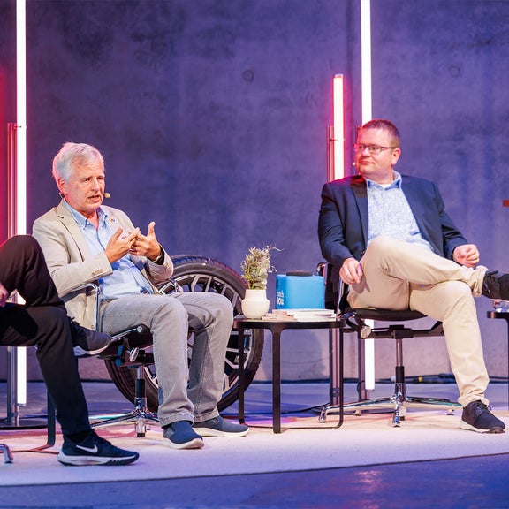 Drei Männer diskutieren auf einer Bühne über Mikroplastik, im Hintergrund leuchten farbige Lichtstäbe.