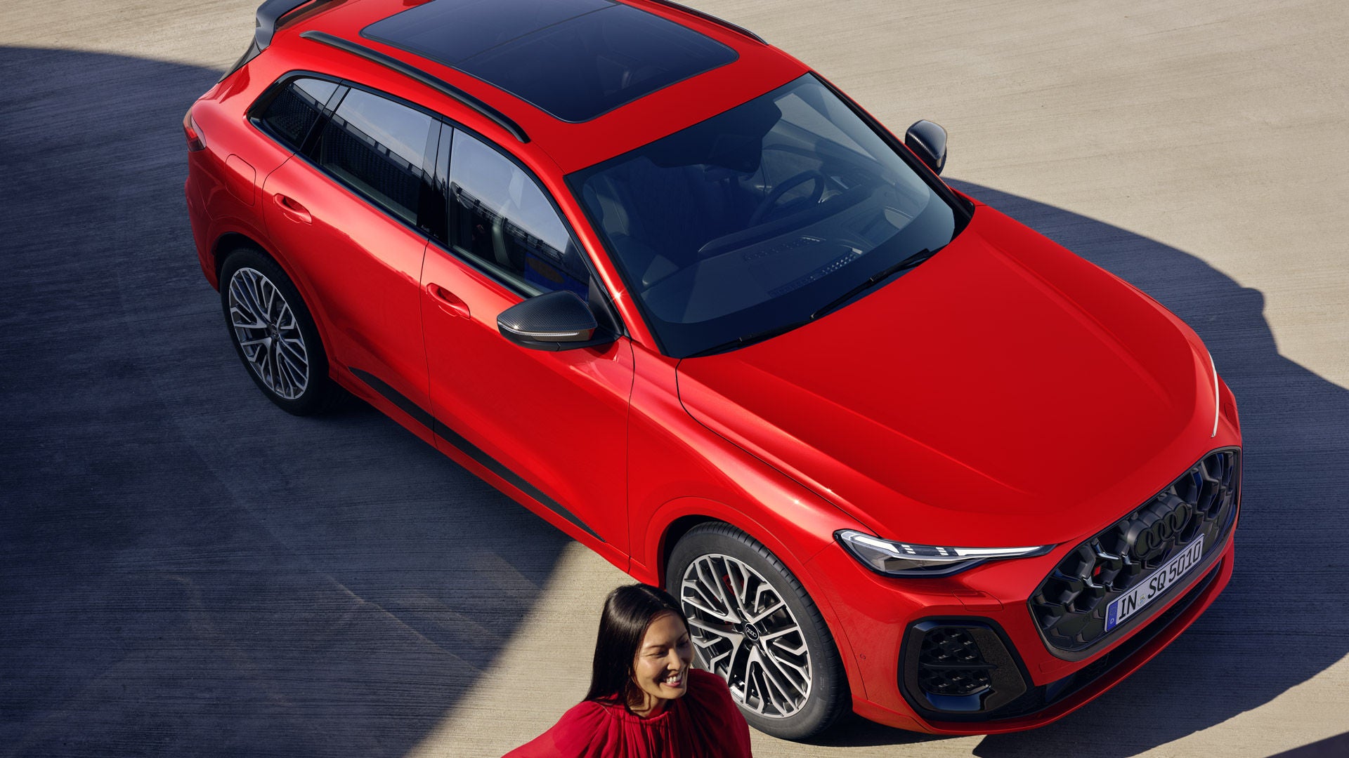 The Q5 SUV viewed from above. The vehicle is painted Misano red. A young woman stands to the right of the front.	