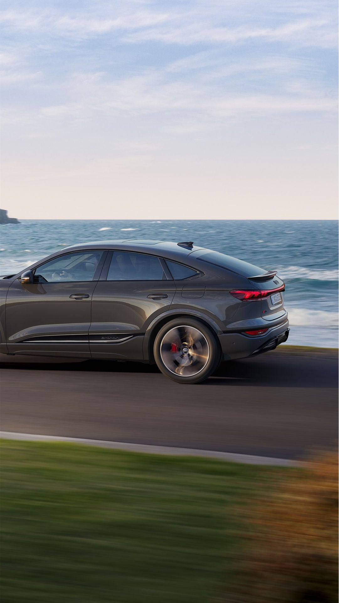 A grey Audi drives along a coastal road with ocean waves and cliffs in the background. The scene conveys a sense of freedom and adventure.