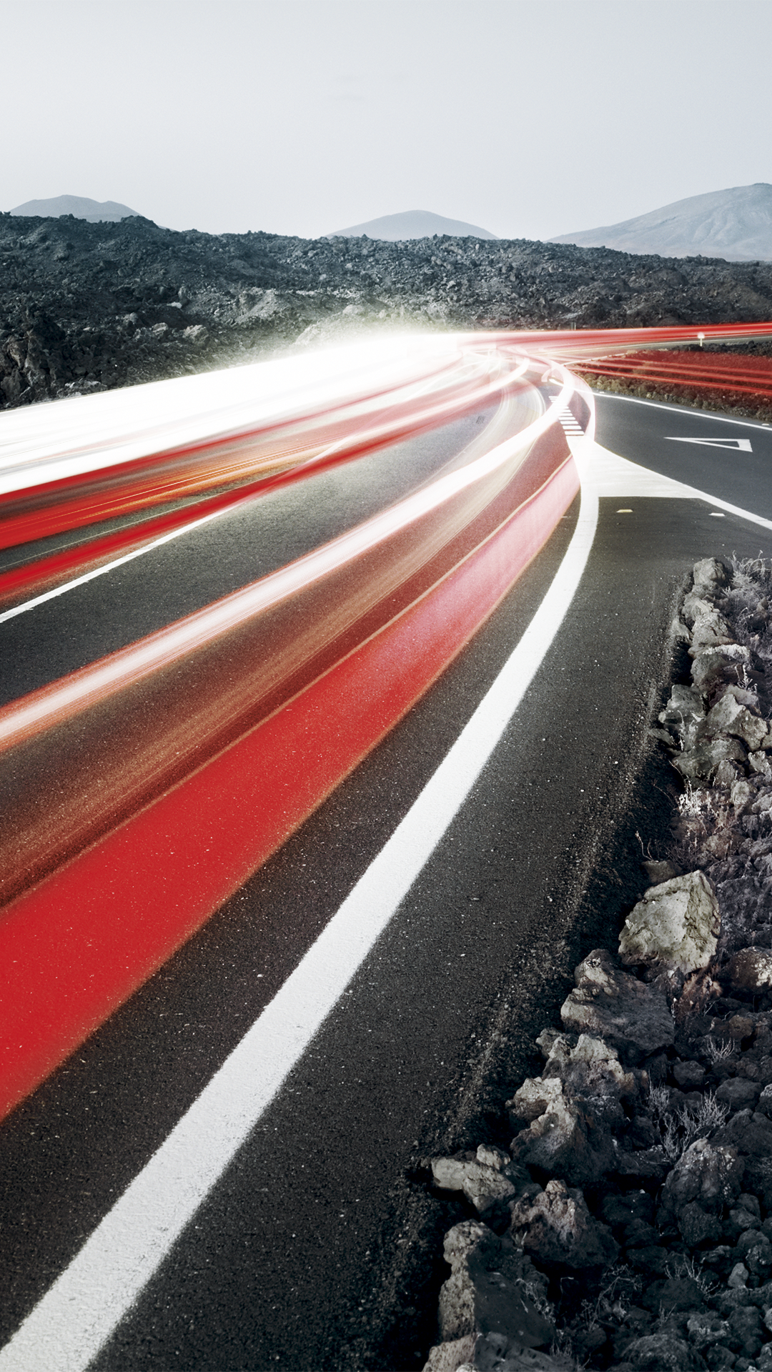 Lichtspuren von Autos auf kurviger Straße durch karge Vulkanlandschaft bei Dämmerung.