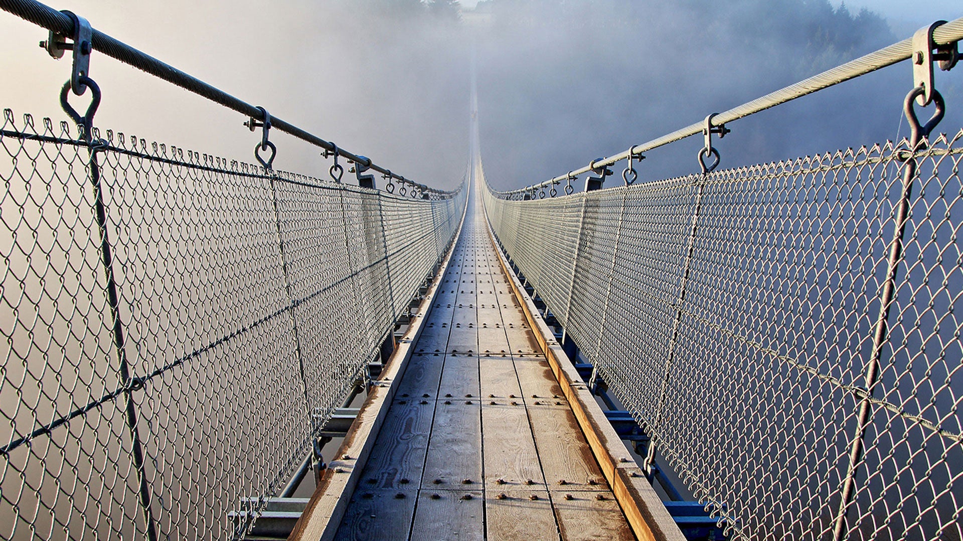 A long suspension bridge with wooden planks and wire ropes leads straight ahead into dense fog over a deep gorge.