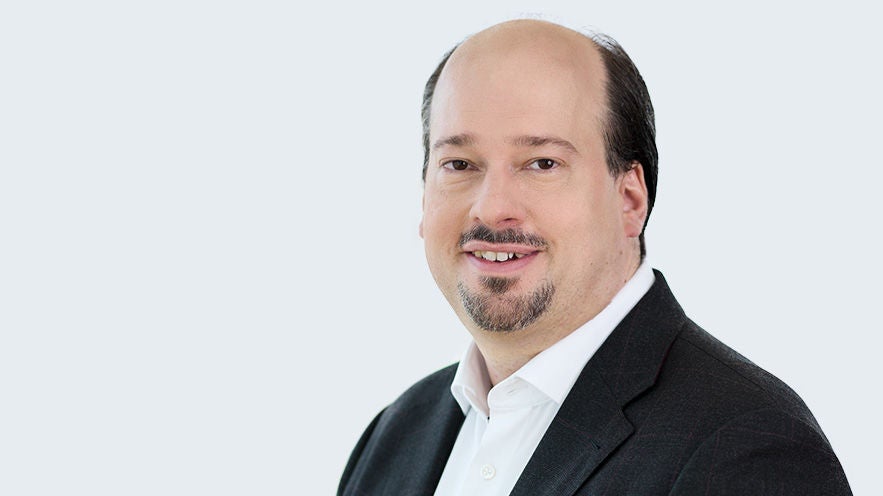 Felix Sedlmaier: Porträt eines lächelnden Mann mit leichtem Bart an Oberlippe und Kinn im Anzug vor grauem Hintergrund.