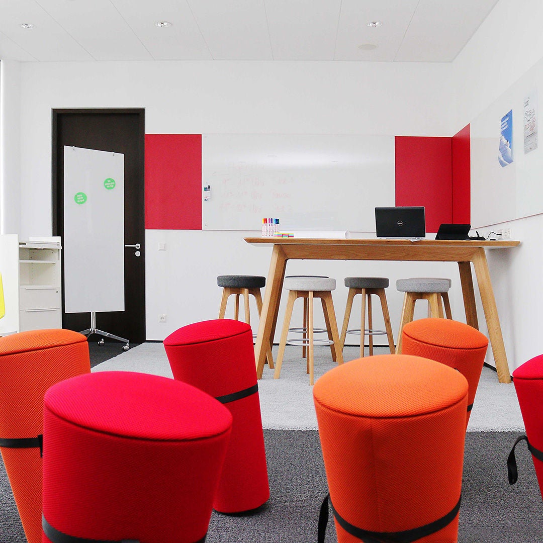 Heller Raum mit hohem Stehtisch, Hockern, bunten Sitzhockern in rot und orange und Whiteboards an rot-weißen Wänden.