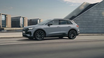 The dark blue Q7 SUV is parked in front of an urban bridge and a mountainous landscape, next to a young, fashionable couple.